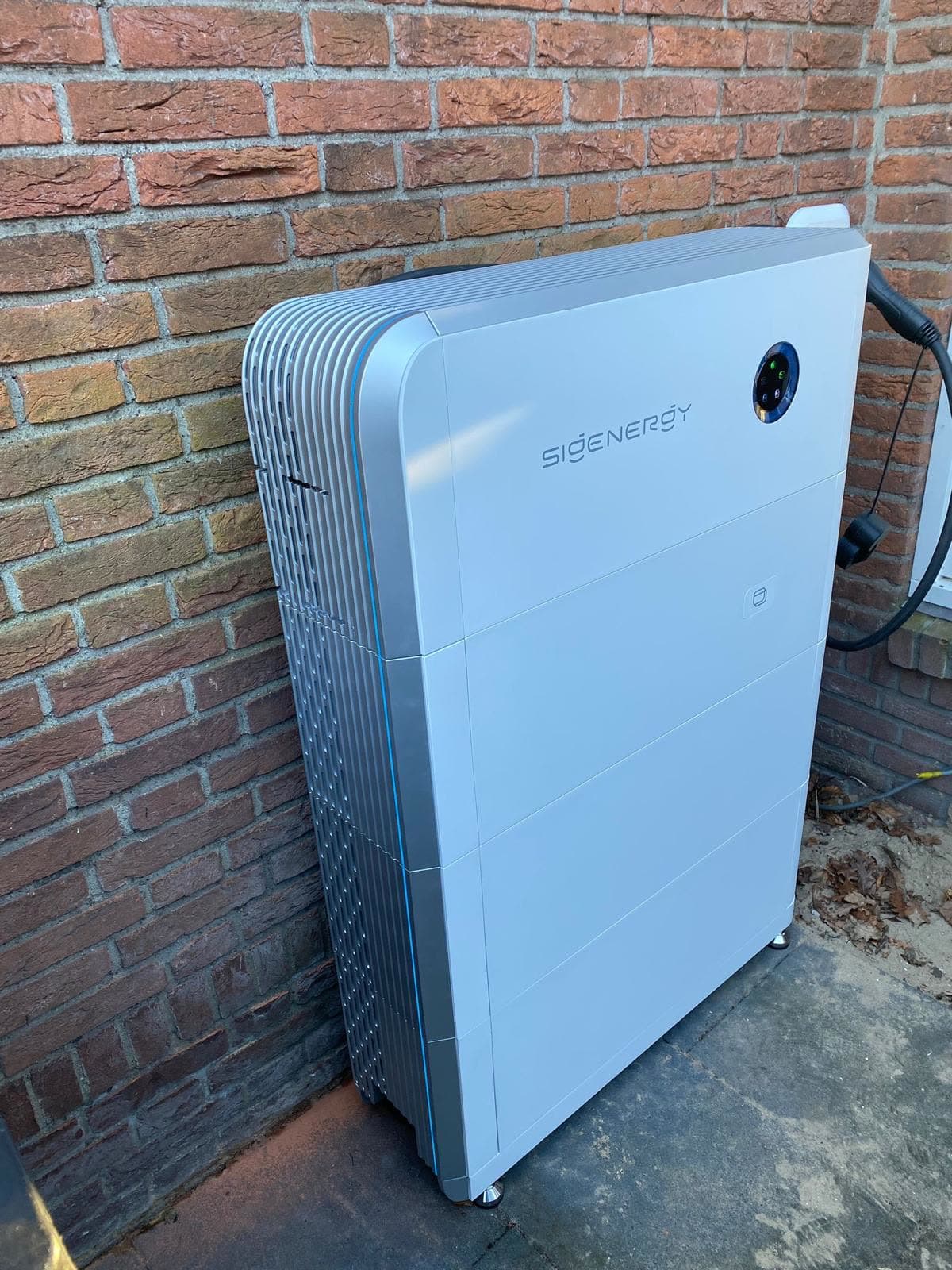 SigenStor-thuisbatterij buiten tegen een bakstenen gevel, met laadkabel aan de zijkant